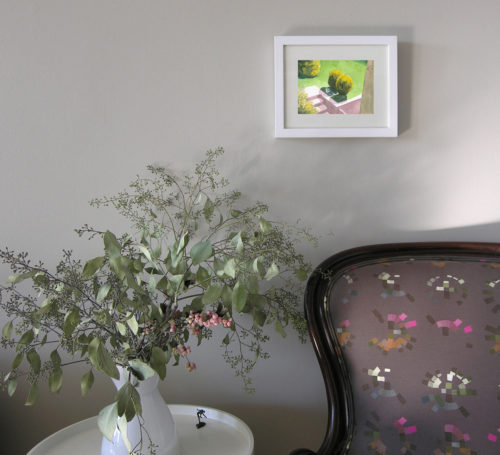 View of a white-framed watercolour painting of a cat napping in the shade of a small bush.