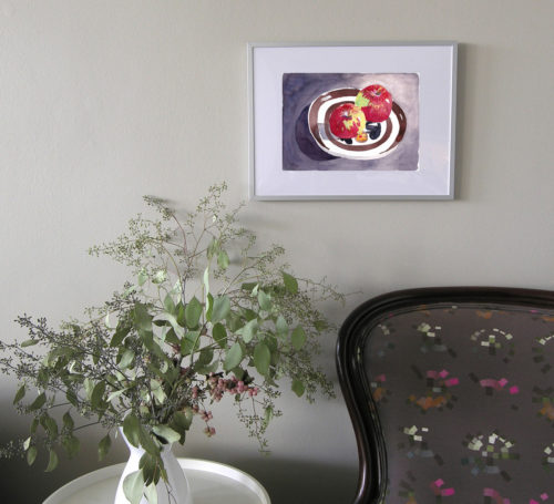 View of a small framed watercolour painting of two red apples on a striped china plate seen on a wall over an armchair