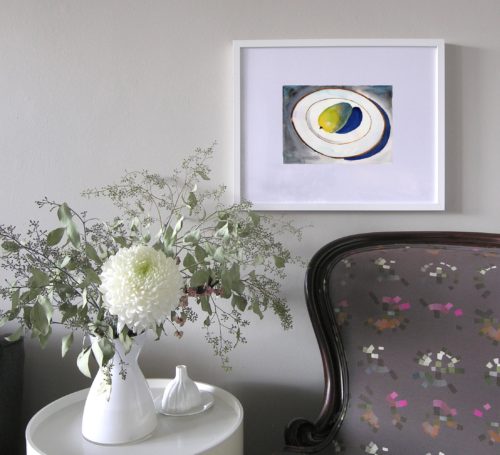 Small framed original watercolour painting of a yellow mango on a white china plate by Michaelle McLean hangs on the wall above an armchair.
