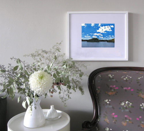 In situ photo of a small watercolour featuring an intense cobalt blue summer sky in a white frame above a bouquet of flowers and an armchair.